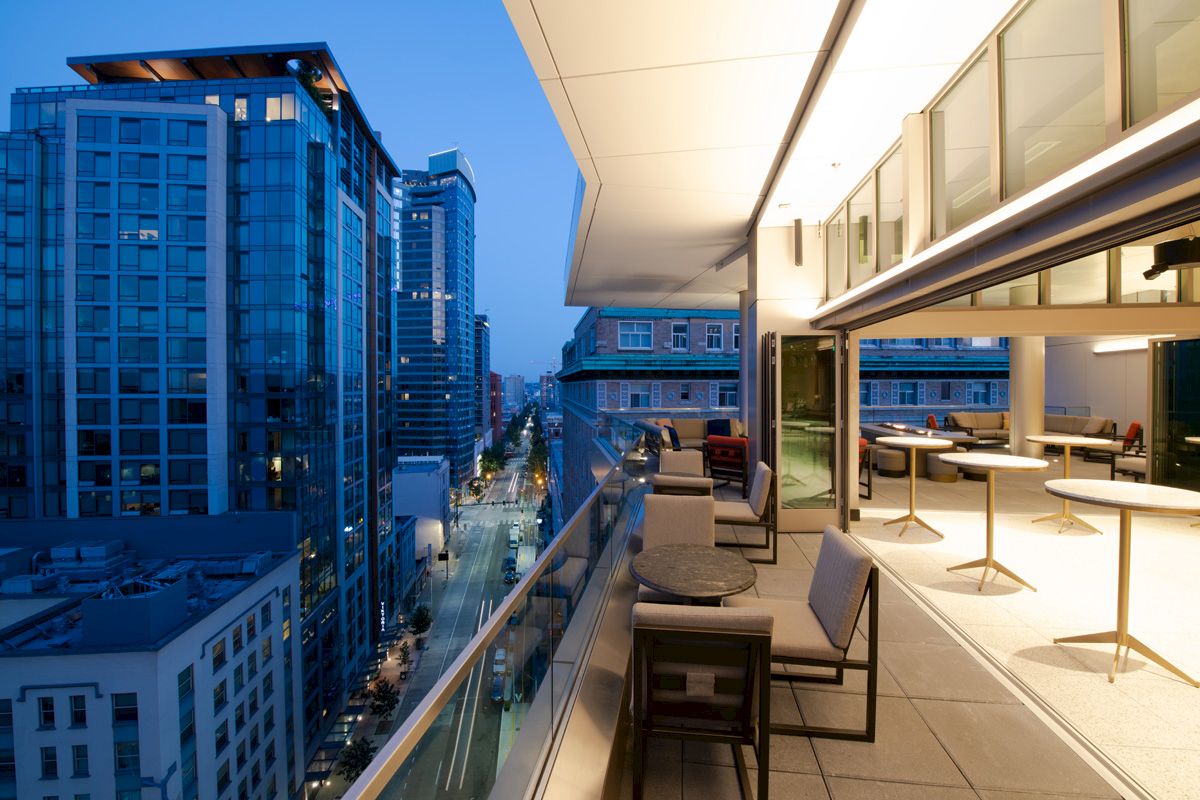 A modern cityscape at dusk featuring a rooftop terrace with seating, overlooking tall buildings and a street below, under a blue sky.