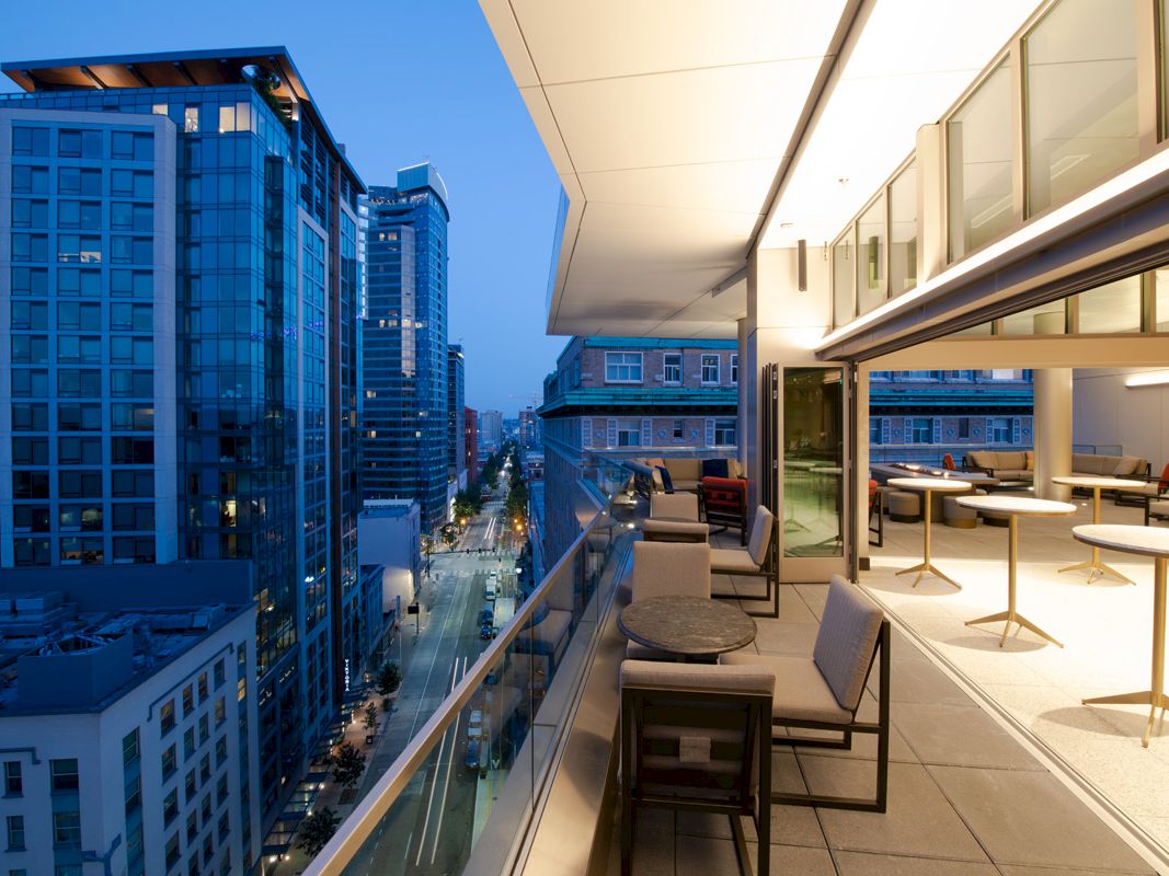 A modern cityscape at dusk featuring a rooftop terrace with seating, overlooking tall buildings and a street below, under a blue sky.