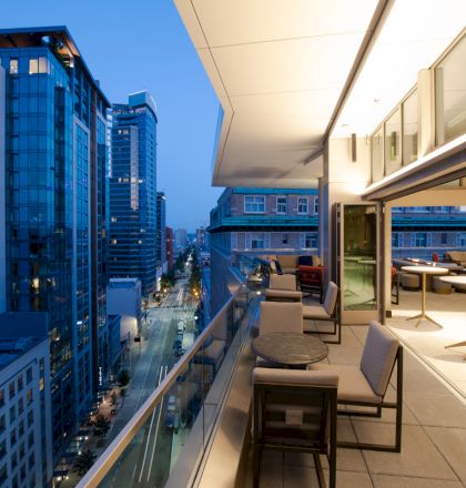 Modern cityscape at dusk with illuminated buildings and a rooftop terrace featuring seating and tables overlooking the street below.