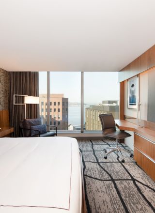 A modern hotel room with a large bed, desk, TV, and a window with a cityscape view. The decor is contemporary and minimalist.