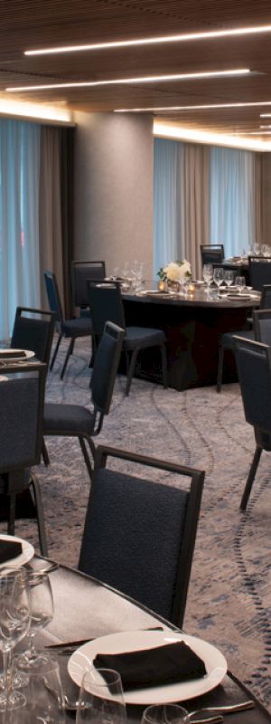 The image shows a modern banquet hall with round tables, chairs, elegant place settings, and soft lighting for an event.