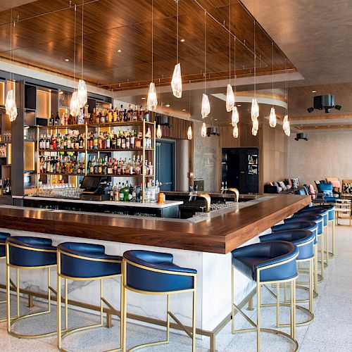 A modern bar with blue stools, a well-stocked liquor display, unique pendant lights, and a stylish seating area in the background.