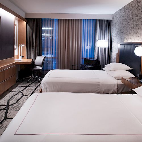 A modern hotel room with two double beds, a wall-mounted TV, desk, chair, and contemporary decor, featuring patterned carpet and wallpaper.