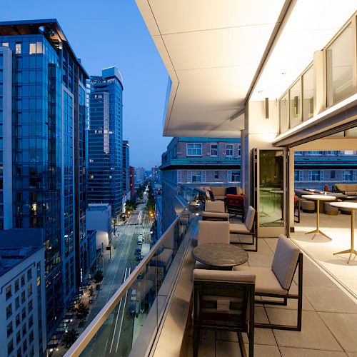 Cityscape view from a modern balcony with outdoor seating, overlooking illuminated buildings and a street at dusk.