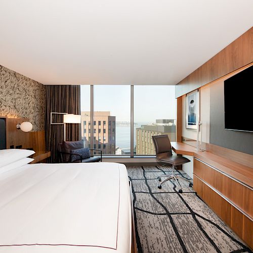 A modern hotel room with a large bed, wall-mounted TV, desk, and chair by a window showing an urban view.