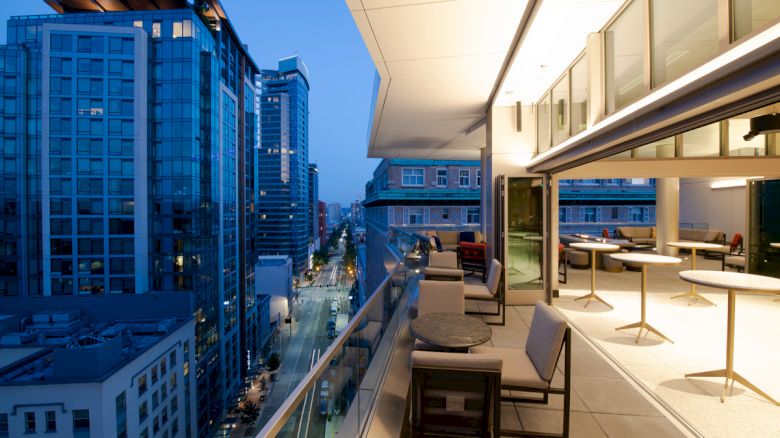 A cityscape at dusk is visible from a modern balcony, featuring tables and chairs, overlooking a street lined with tall buildings.