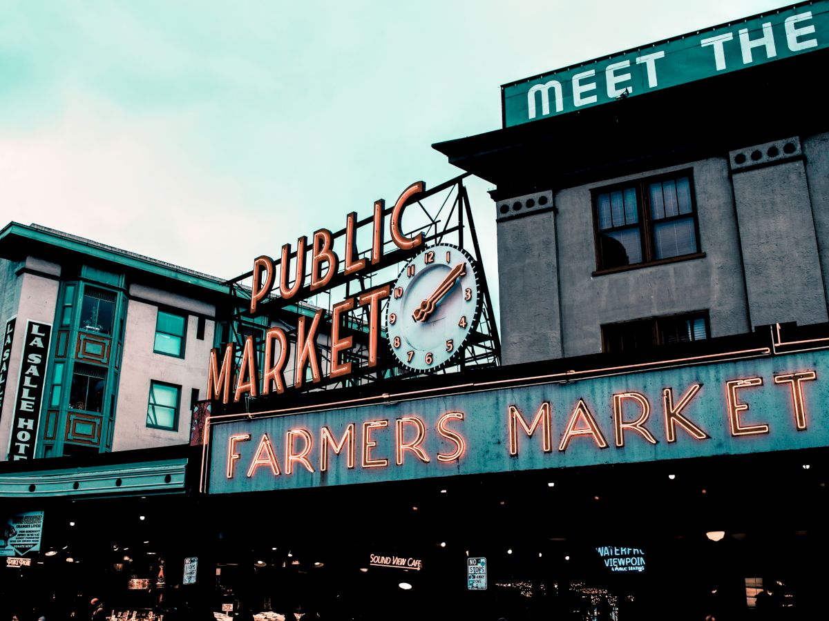 The image shows a famous market sign, "PUBLIC MARKET" and "FARMERS MARKET," with buildings in the background.