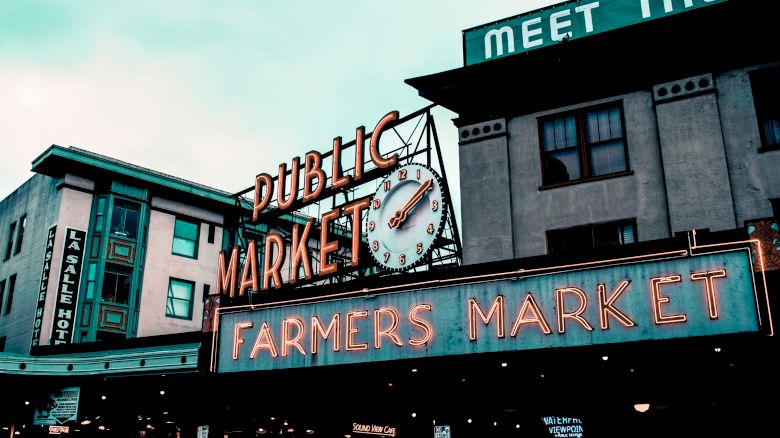 The image shows a famous market sign, "PUBLIC MARKET" and "FARMERS MARKET," with buildings in the background.