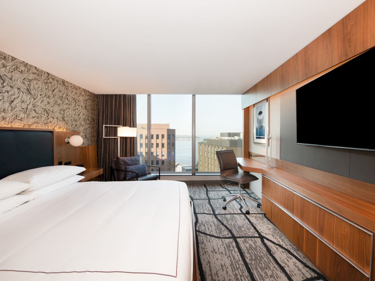 A modern hotel room with a bed, large TV, desk, and city view through floor-to-ceiling windows, featuring a contemporary design aesthetic.
