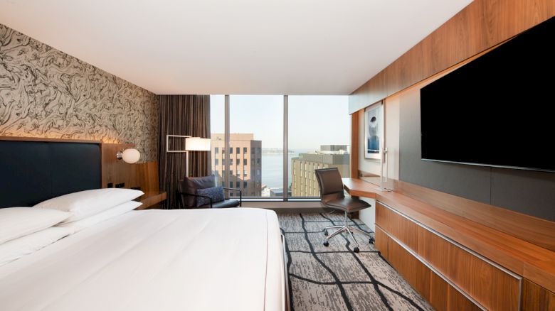 A modern hotel room with a bed, large TV, desk, and city view through floor-to-ceiling windows, featuring a contemporary design aesthetic.