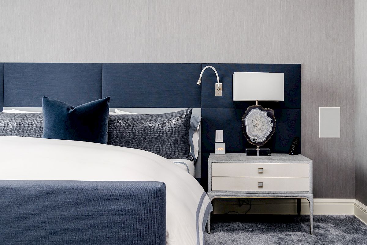 A modern bedroom with a blue bed, white and gray linens, a nightstand, lamp, and wall-mounted reading light.