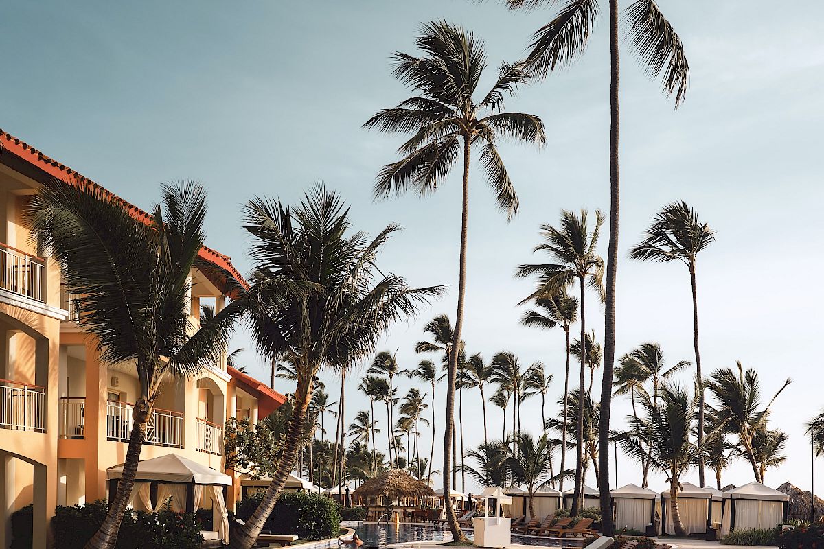 A resort pool lined with lounge chairs and palm trees beside a building under a blue sky, creating a tropical atmosphere.