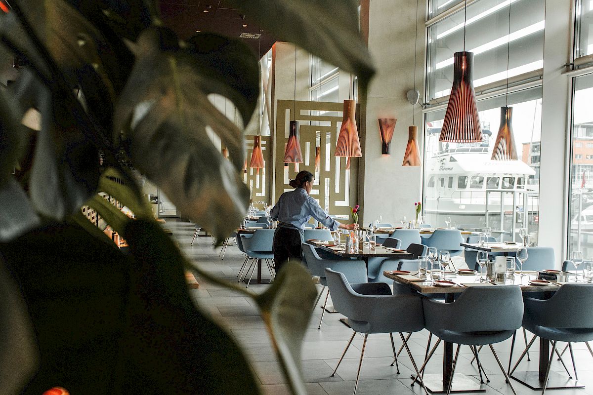 A person is setting tables in a modern restaurant with hanging lights and plants visible in the foreground.