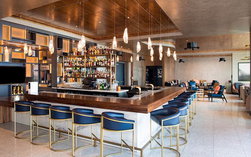 A modern bar and lounge area with blue barstools, hanging pendant lights, a stocked bar, and comfortable seating in the background.