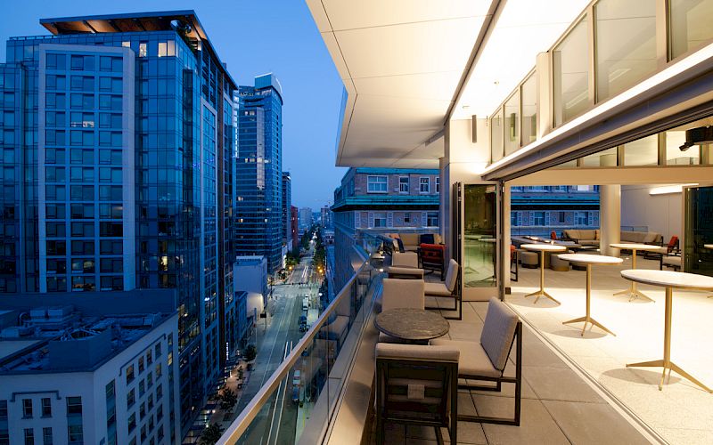 The image shows a modern outdoor terrace with seating, overlooking a city street with tall buildings during twilight.