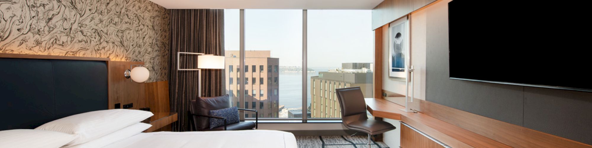 A modern hotel room features a bed, desk, large TV, and window with a city view, creating a sleek and comfortable space.