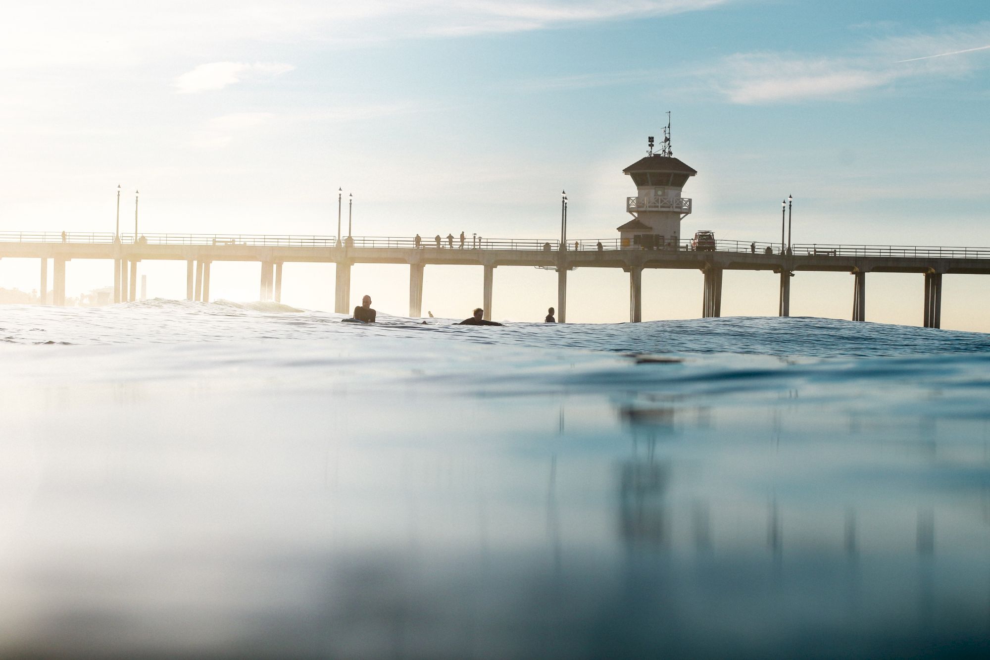 A pier extends over the ocean with people on it, while surfers are visible in the water near the foreground of the image.